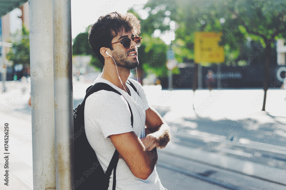 Young man with dark curly hair wearing sunglasses is smiling at the camera while standing on the street with a backpack on his back. Young bearded male is waiting for a transport on a station.