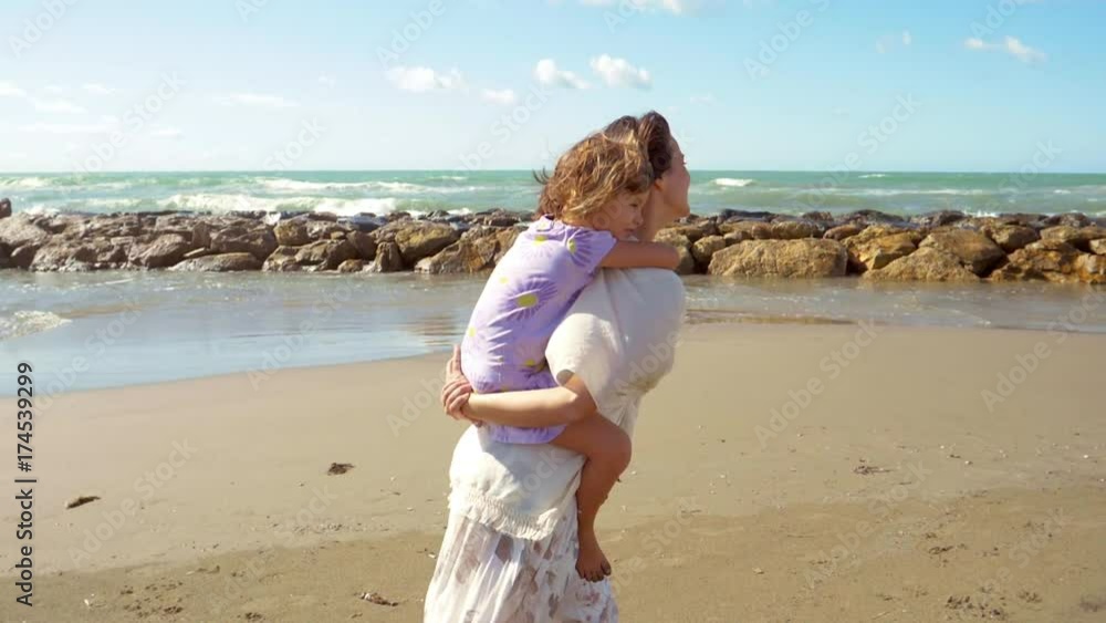 Woman walking on beach holding little daughter on back smiling talking in front of ocean happy Woman walking on beach holding little daughter on back smiling talking in front of ocean happy