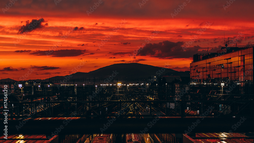 Fototapeta premium Stunning sunset on huge railroad terminal with multiple railway tracks, glass facade on the right and hills ridge in distance; reddish sky, autumn evening, many reflections and lights, Adler, Russia