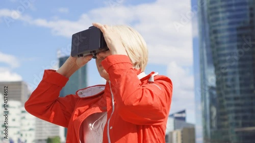 Senior woman in red coat standing in downtown business dictrict using VR glasses. Skyscrapers on background photo