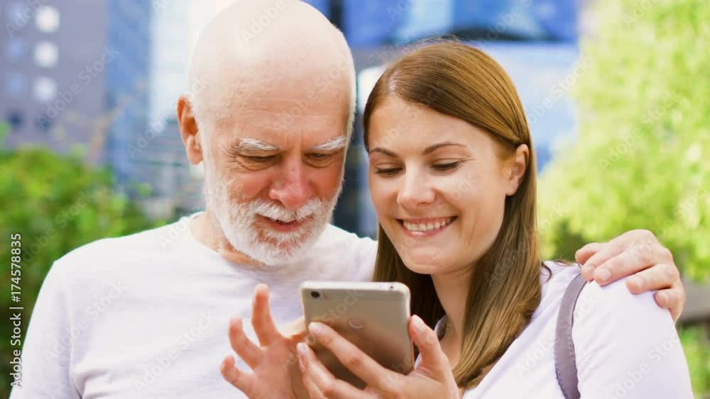 Senior father and his young daughter standing on street using mobile phone. Happy family enjoying time together. Daughter teaching father how to use cellphone Senior father and his young daughter standing on street using mobile phone. Happy family enjoying time together. Daughter teaching father how to use cellphone