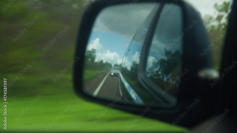 A close up shot of a car's side mirror reflecting the bad weather and a car behind the vehicle. Green grass are seen beside the road. A close up shot of a car's side mirror reflecting the bad weather and a car behind the vehicle. Green grass are seen beside the road.