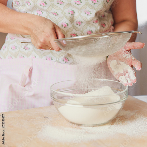 Fotografija Aged female hands sifting flour by sieve in glass bowl.