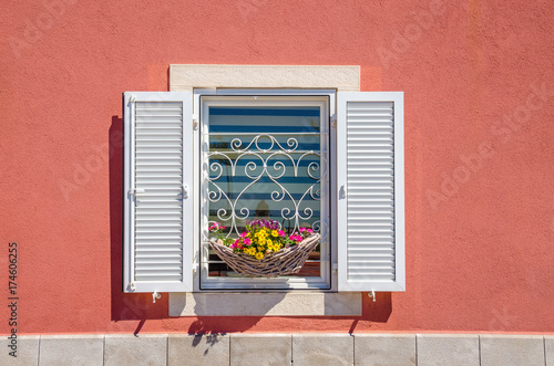 Window with white shutters and beautiful blooming flowers against a red wall
