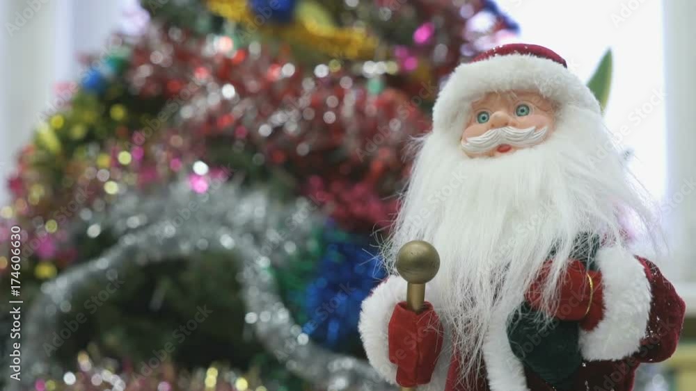 Close up of toy Santa Claus standing next to the christmas tree and slowly moves his hands in different directions in kindergarten at the New Year and Christmas holiday. Close up of toy Santa Claus standing next to the christmas tree and slowly moves his hands in different directions in kindergarten at the New Year and Christmas holiday.