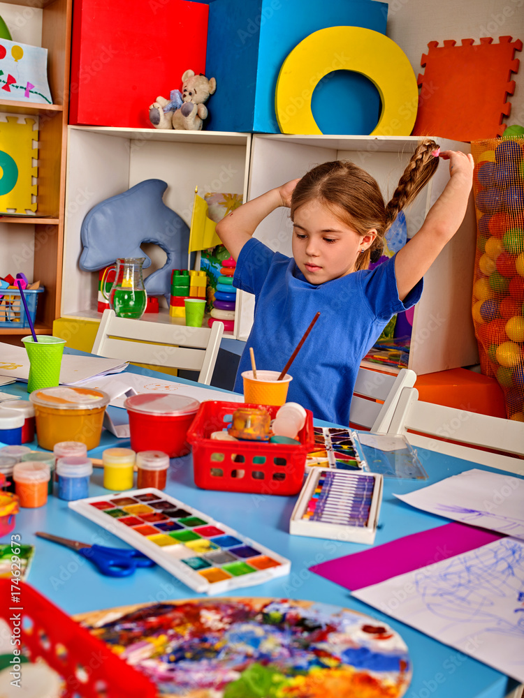 Fototapeta premium Small student girl painting in art school class. Child drawing by paints on table. Girl in an orphanage. Craft drawing education develops creative abilities of children. Children indulge.