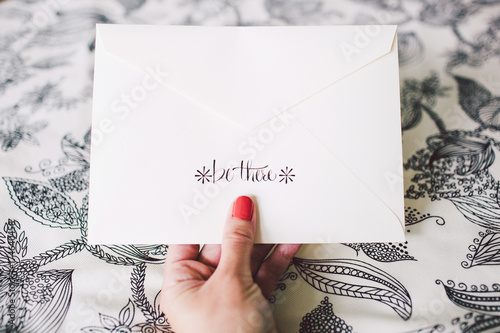 A hand holding an invitation envelope with hand lettering that readsbe there 