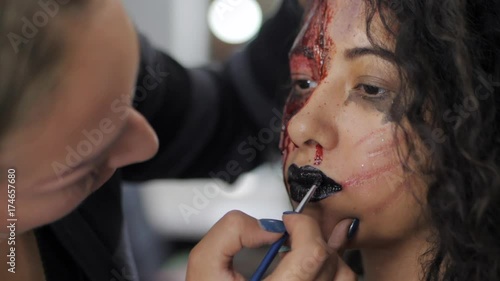 Make-up artist make the girl halloween make upin studio.Halloween face art. Woman applies on black lipstick with brush on lips of latin girl. War-paint with blood, scars and wounds.Slow motion. photo