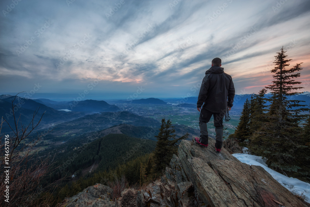 Man watching a beautiful cloudy sunset over a rocky cliff. Picture taken on top of Elk Mountain near Chilliwack, British Columbia, Canada.