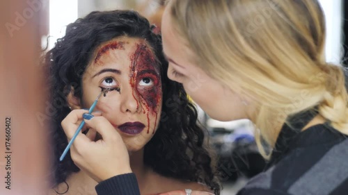 Make-up artist make the girl halloween make upin studio.Halloween face art.Woman applies on professional greasepaint on the face of spanish girl.War-paint with blood, scars and wounds. 4k photo