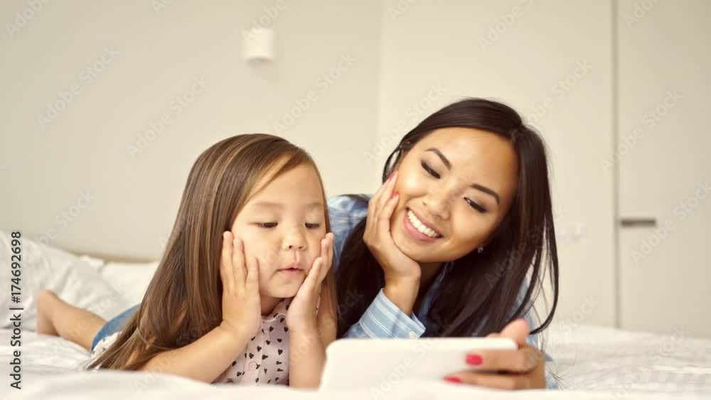 Smiling pretty asian woman lying on bed with her young daughter and using smartphone Smiling pretty asian woman lying on bed with her young daughter and using smartphone