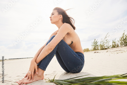 An asian girl topless sitting on a beach on sand in backlight with a bunch of grass dreaming