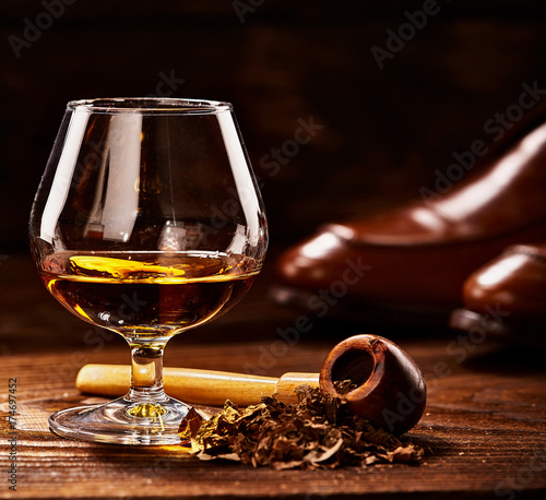 Glass of cognac and pipe with tobacco with glance shoes defocused on second background on wooden table photo