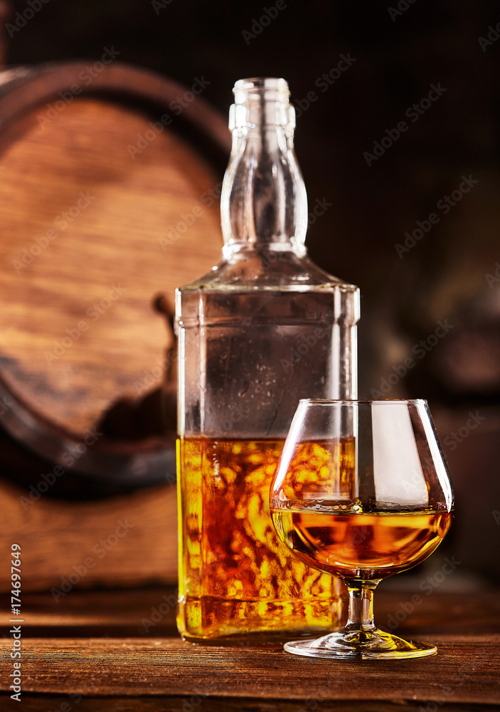 Glass and bottle of Cognac on wodden table old oak barrel defocussed Glass and bottle of Cognac on wodden table old oak barrel defocussed