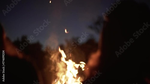 silhouettes of two girls sitting in front bonfire with flame sparks and smoke rising to sky near wood slow motion closeup