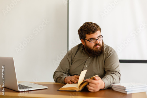 Pensive young european businessman reading book at desktop with laptop, Knowledge concept