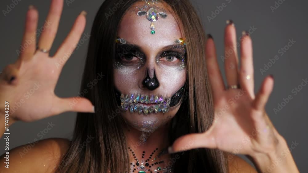 Young girl with creative halloween face art frighten in the studio. Scary portrait of glamorous skull with rhinestones and sequins. Professional make-up for the celebration. Slow motion. Young girl with creative halloween face art frighten in the studio. Scary portrait of glamorous skull with rhinestones and sequins. Professional make-up for the celebration. Slow motion.
