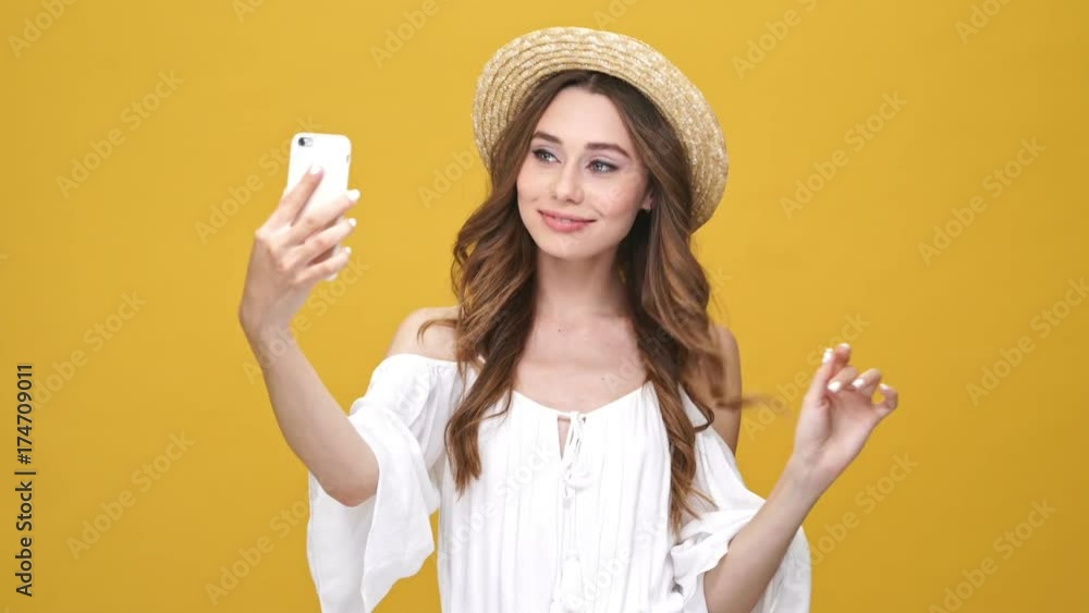 Beauty playful brunette woman in dress and hat posing and making selfie on her smartphone over orange background Beauty playful brunette woman in dress and hat posing and making selfie on her smartphone over orange background