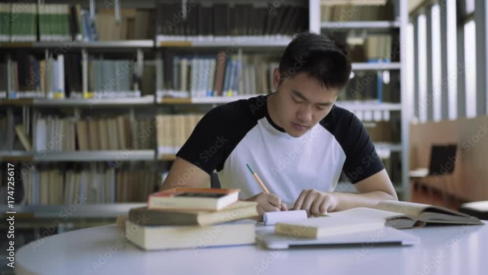 Tracking shot of student studying at desk in library in college. A high school student writes essay in library. Book shelves in background Tracking shot of student studying at desk in library in college. A high school student writes essay in library. Book shelves in background