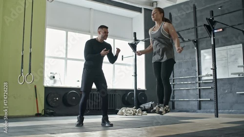 Personal coach checking time on sports timer and motivating young woman while she jumping the rope in cross training gym