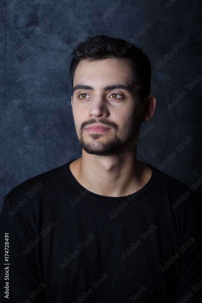 portrait of a young man on a dark background. A brunette, a white man, dressed in all dark. short beard and mustache. Emotions.