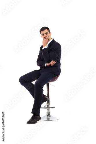 A young man sits on a bar high chair, a business suit, a serious look, is inexhaustible or a financier, or an office employee. Isolate on white background.
