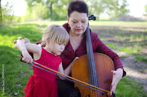Old women and blond caucasian girl playing concert on cello outdoors. Happy grandmother teaching to play violoncello her granddaughter.