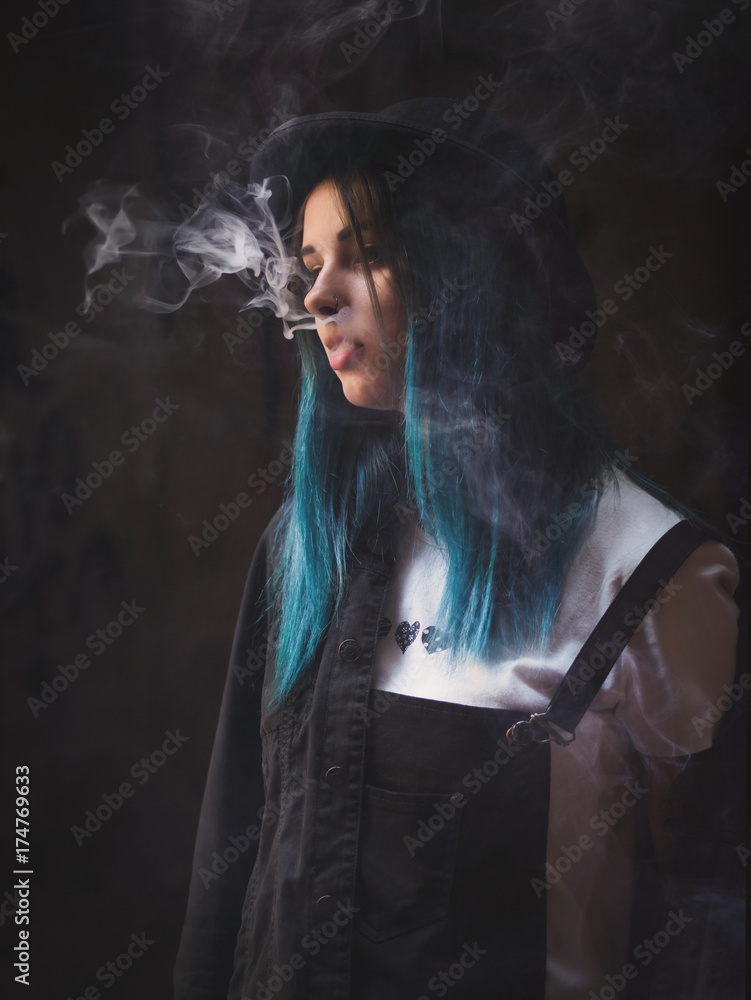 Emo girl smoking  punk or hipster woman with blue colorful  dyed hair, hat, piercing,lenses,ears tunnels and unusual hairstyle stands  in backyard. beautiful smoke. Stock Photo | Adobe Stock