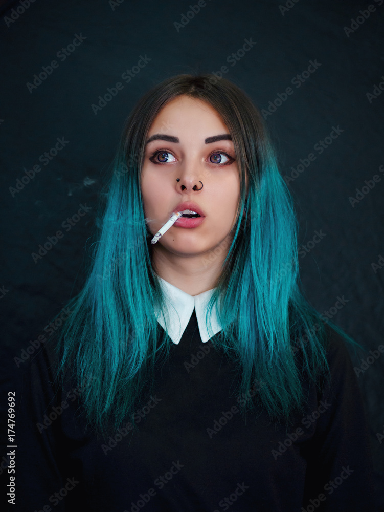 Emo girl smoking cigarette. Young student or pupil with blue colorful dyed  hair, hat, piercing,lenses,ears tunnels and unusual hairstyle stands on  black background. Stock Photo