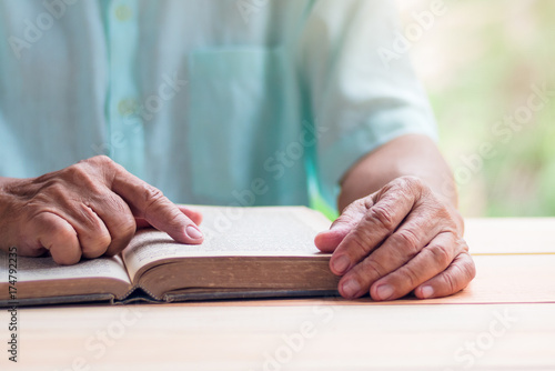 old man reading book on light brown wooden table surface, pointing with right index finger, selective focus photo