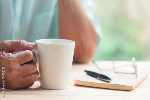 old man hold white cup of tea or coffee with right hand on light brown wooden table surface, blurred pen, notebook and glasses as background