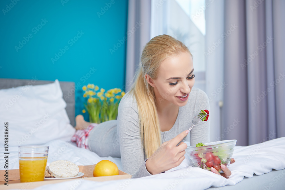 Smiling young blond woman lying in the bed and eating a green salad Smiling young blond woman lying in the bed and eating a green salad