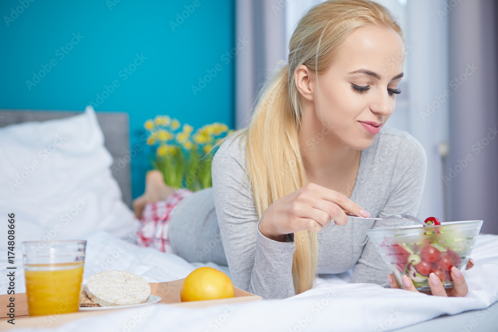 Smiling young blond woman lying in the bed and eating a green salad