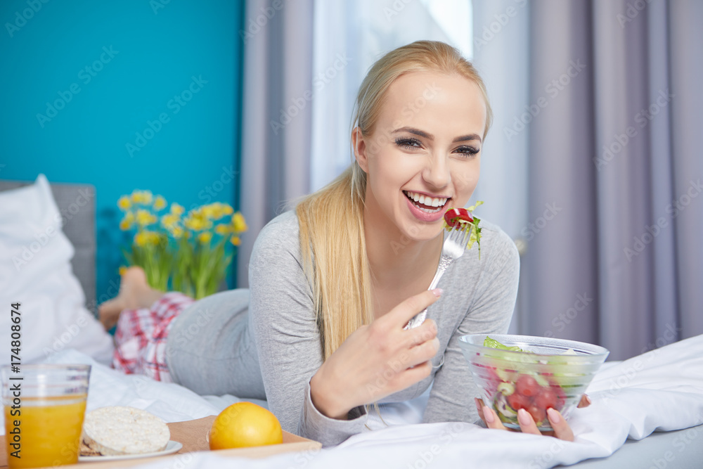 Smiling young blond woman lying in the bed and eating a green salad Smiling young blond woman lying in the bed and eating a green salad
