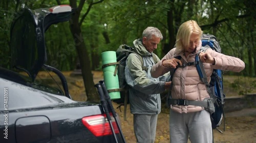 Nice elderly man helping his wife witrh wearing a tourism backpack