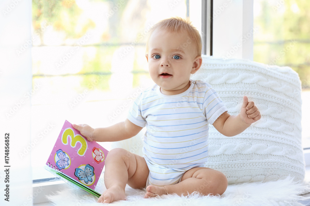 Cute little baby with book sitting on window sill at home Cute little baby with book sitting on window sill at home