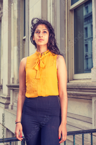 East Indian American college student with long curly hair studying in New York, wearing sleeveless orange shirt, striped pants, standing by vintage style office building. Instagram filtered effect..