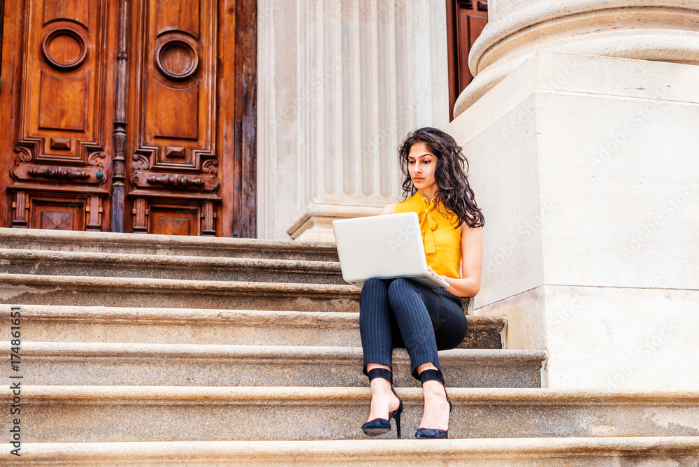 East Indian American college student studying in New York, wearing sleeveless orange shirt, striped pants, high heels, sitting on stairs outside office building on campus, working on laptop computer.