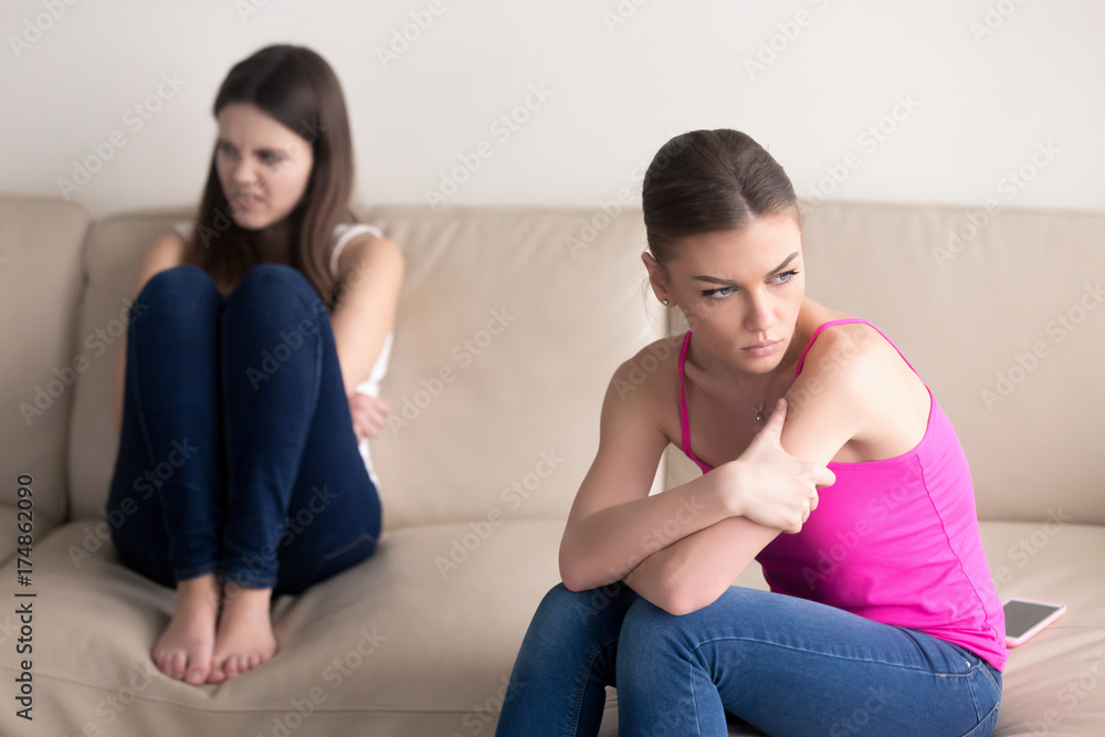 Girlfriends sitting on sofa, hating each other after heated argument. Girl on foreground is very upset, her sister has hateful facial expression. Friends having bad argument, crisis in relationship. Girlfriends sitting on sofa, hating each other after heated argument. Girl on foreground is very upset, her sister has hateful facial expression. Friends having bad argument, crisis in relationship.