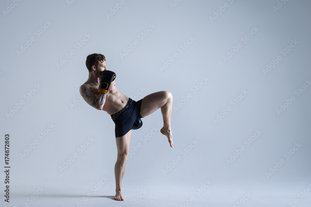 Horizontal picture of mixed fight man in trunks and gloves practising kicks in spacious studio. Attractive professional male kickboxer training in gym. Sports, endurance, determination and activity Horizontal picture of mixed fight man in trunks and gloves practising kicks in spacious studio. Attractive professional male kickboxer training in gym. Sports, endurance, determination and activity