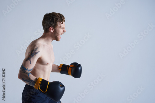 Принти на полотні Profile shot of fearless young male boxer with athletic body screaming during fight, having firce aggressive look, getting mad at his opponent because of foul blows
