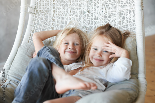Принти на полотні Picture of two siblings spending time together: blonde cute boy sitting in wicker hanging chair with his adorable little sister, both children looking at camera and smiling