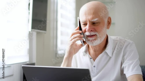 Good-looking handsome senior man sitting at home with laptop and smartphone. Using cellphone discussing project on screen. Remote freelance work on retirement, active modern lifestyle of older people