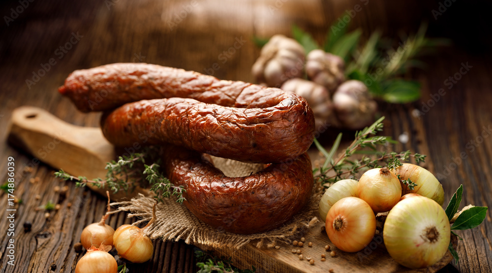 Smoked  sausage on a wooden rustic table with addition of fresh aromatic herbs and spices, natural product from organic farm, produced by traditional methods - obrazy, fototapety, plakaty 