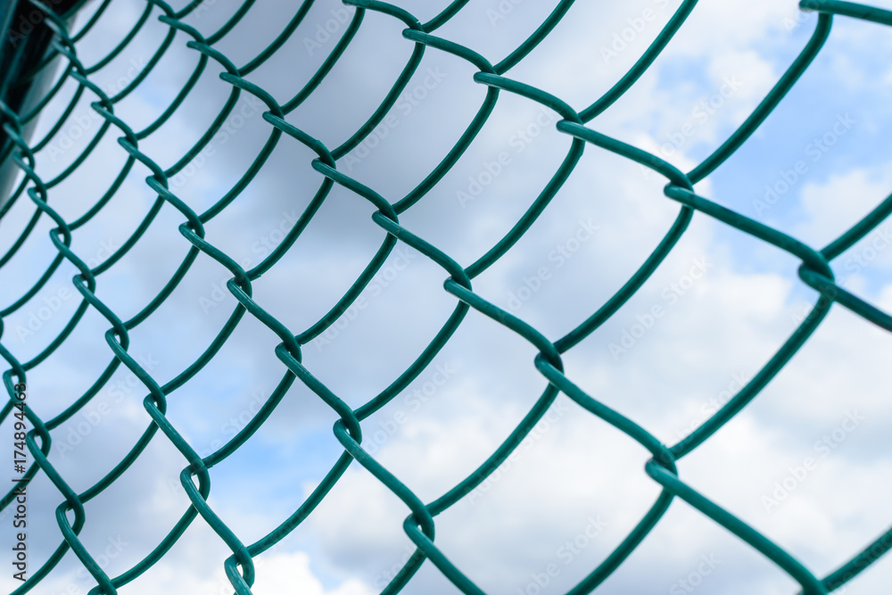 Fototapeta premium Green metal wire fence under fluffy white clouds and blue sky background.