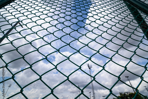 Green metal wire fence under fluffy white clouds and blue sky background.