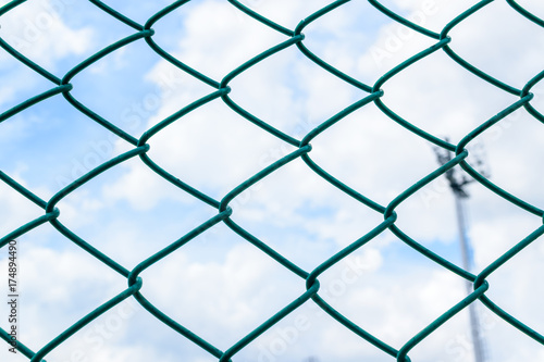 Green metal wire fence under fluffy white clouds and blue sky background.