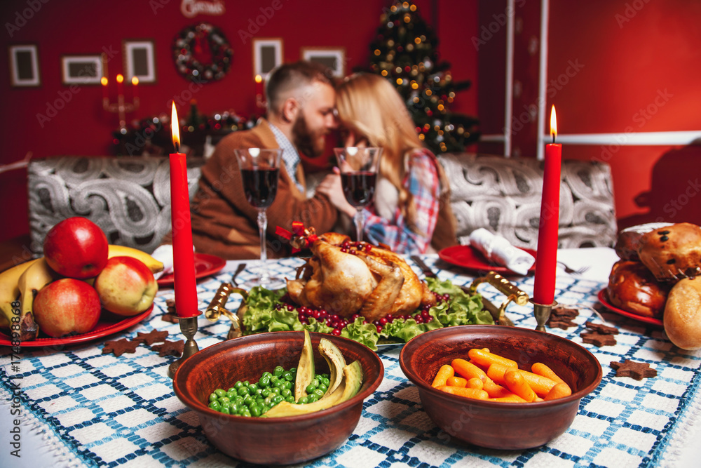 Beautiful couple kissing and holding glass of wine in a decorated festive interior with a Christmas tree. A romantic dinner for thanksgiving with fried chicken and candles