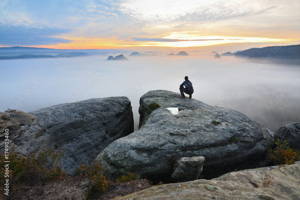 Hiker in squatting position on peak of rock and watching into colorful mist and fog in morning valley.