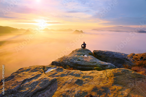 Hiker in squatting position on peak of rock and watching into colorful mist and fog in morning valley.